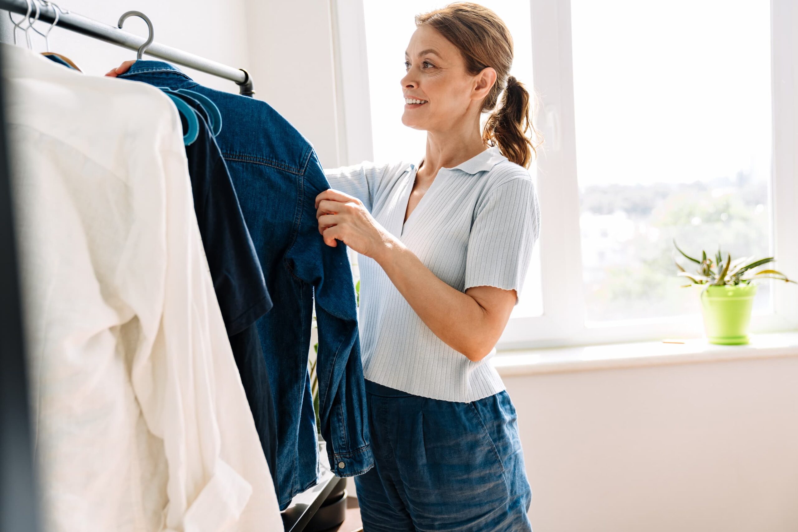 Eine Frau steht in einem hellen, sonnendurchfluteten Raum vor einer Kleiderstange und betrachtet bzw. sortiert Kleidung. Sie hält ein blaues Kleidungsstück in den Händen, während daneben weitere Kleidungsstücke wie Hemden und Jeans hängen. Die Frau lächelt leicht und wirkt zufrieden beim Auswählen ihres Outfits. Im Hintergrund ist ein grosses Fenster mit viel Tageslicht sowie eine kleine grüne Pflanze auf der Fensterbank zu sehen.