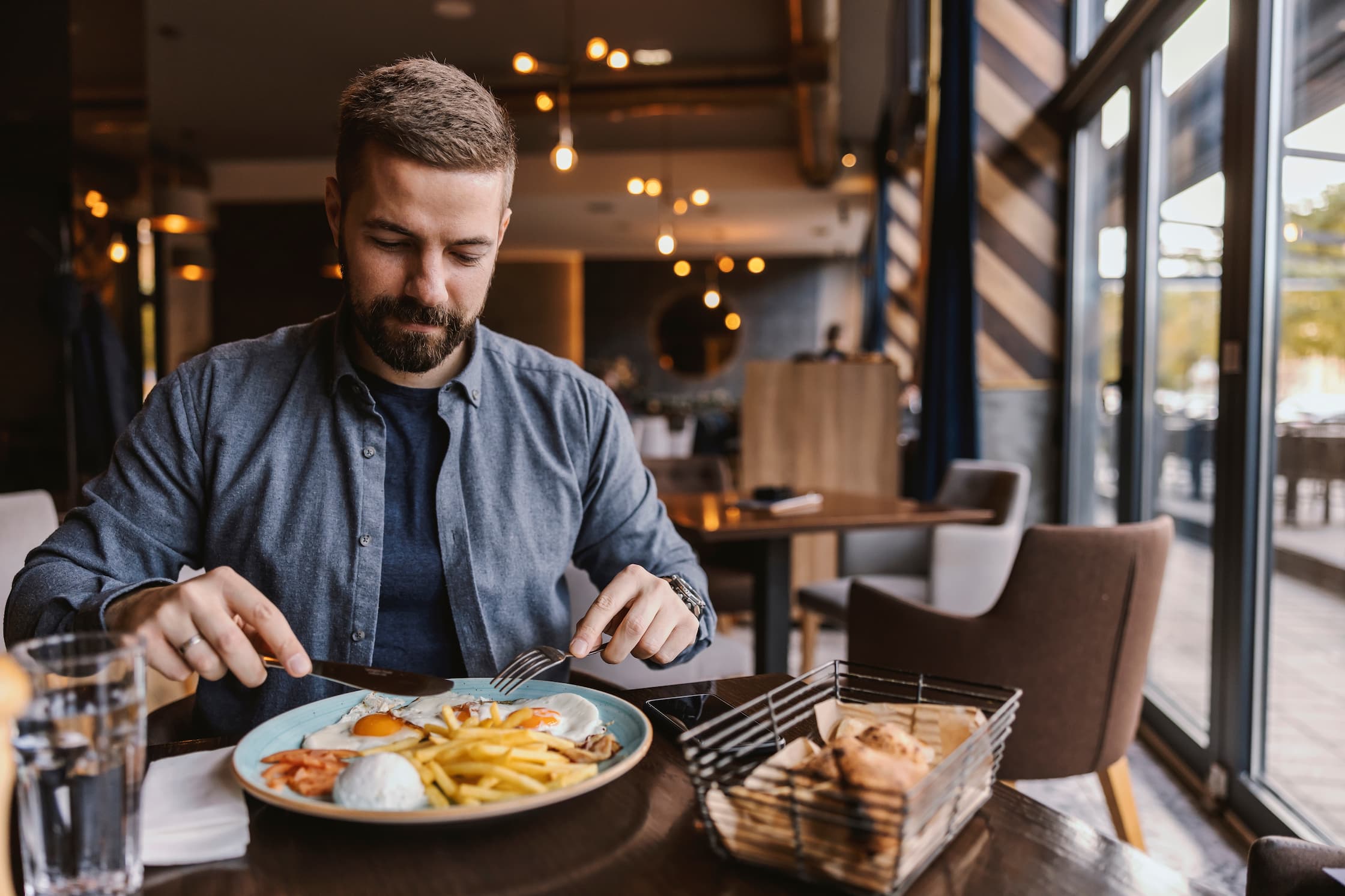 Ein Mann mit Bart sitzt in einem gemütlichen, modern beleuchteten Restaurant und isst eine Mahlzeit aus Spiegeleiern, Pommes und Beilagen von einem Teller, während er mit Messer und Gabel schneidet; auf dem Tisch stehen ausserdem ein Getränk und ein Brotkorb, im Hintergrund sind warme Lichter und weitere Sitzplätze zu sehen.