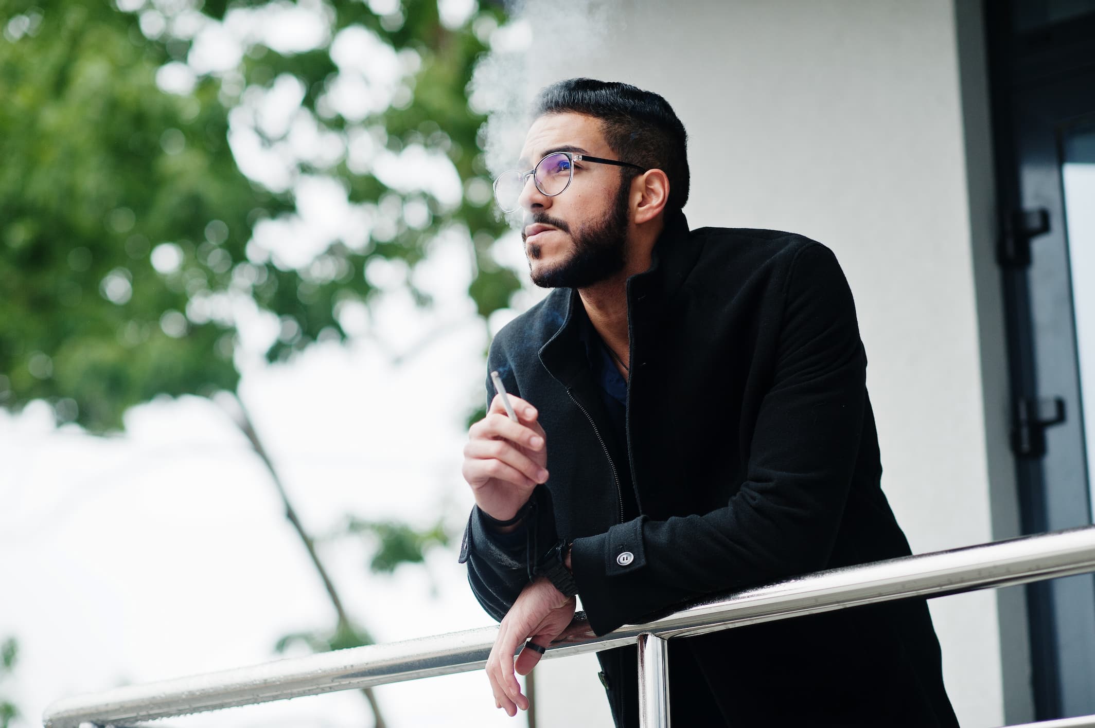 Ein junger Mann mit Brille und kurzem Bart lehnt an einem Geländer im Freien und raucht eine Zigarette. Er trägt einen dunklen Mantel und blickt nachdenklich in die Ferne. Im Hintergrund sind unscharf grüne Bäume und eine helle Gebäudewand zu sehen.