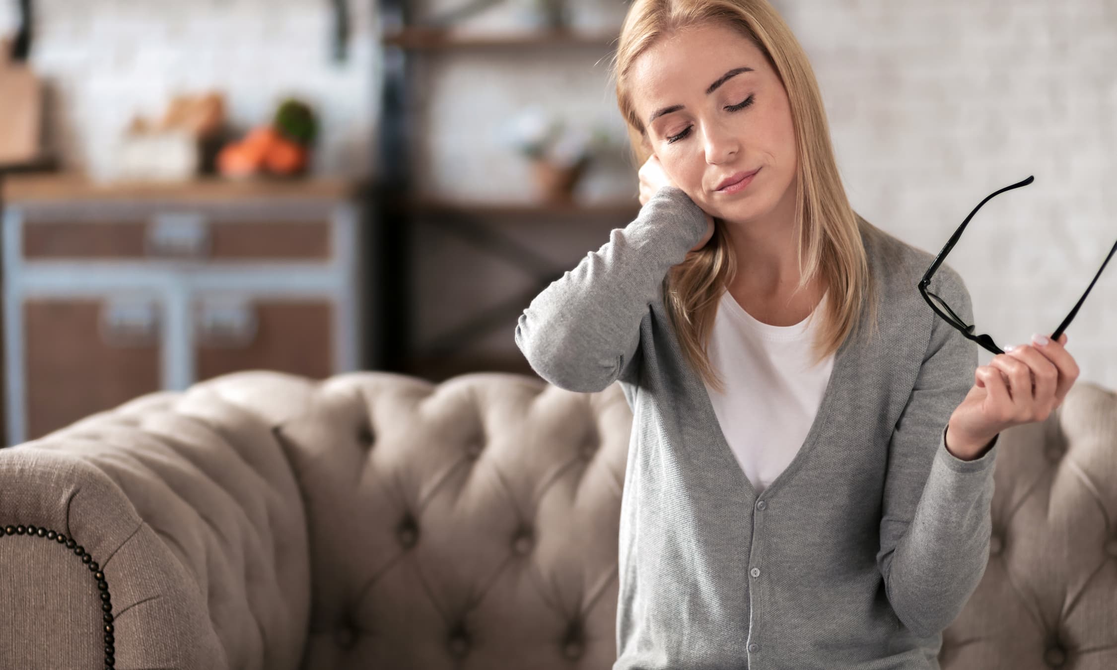 Junge Frau sitzt auf einem Sofa in einem Wohnzimmer, hält eine Brille in der Hand und massiert sich mit geschlossenen Augen den Nacken, als ob sie Verspannungen oder Müdigkeit spürt.