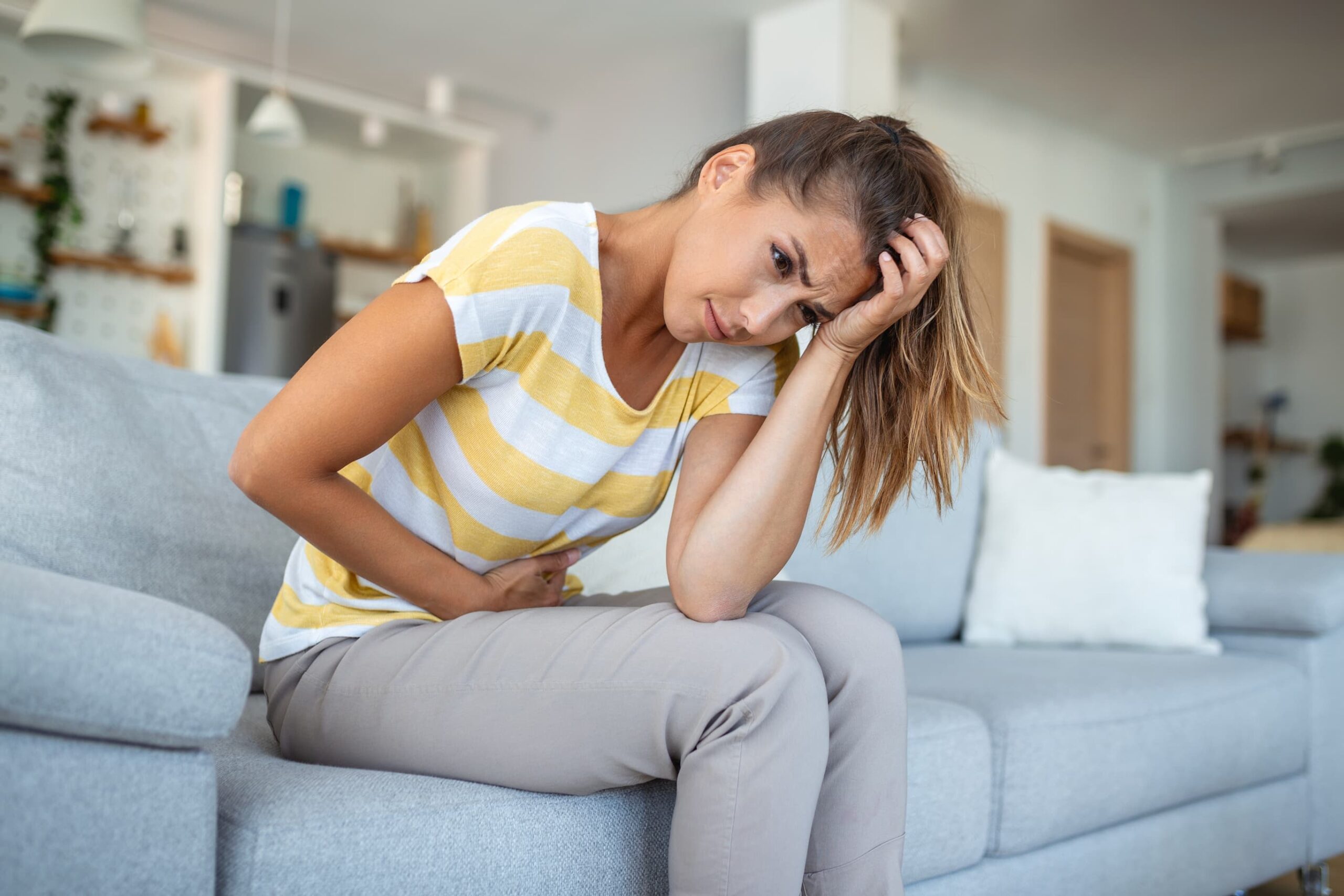 Frau sitzt auf einem Sofa, hält sich den Bauch und den Kopf und wirkt erschöpft oder hat Schmerzen im Bauchbereich.