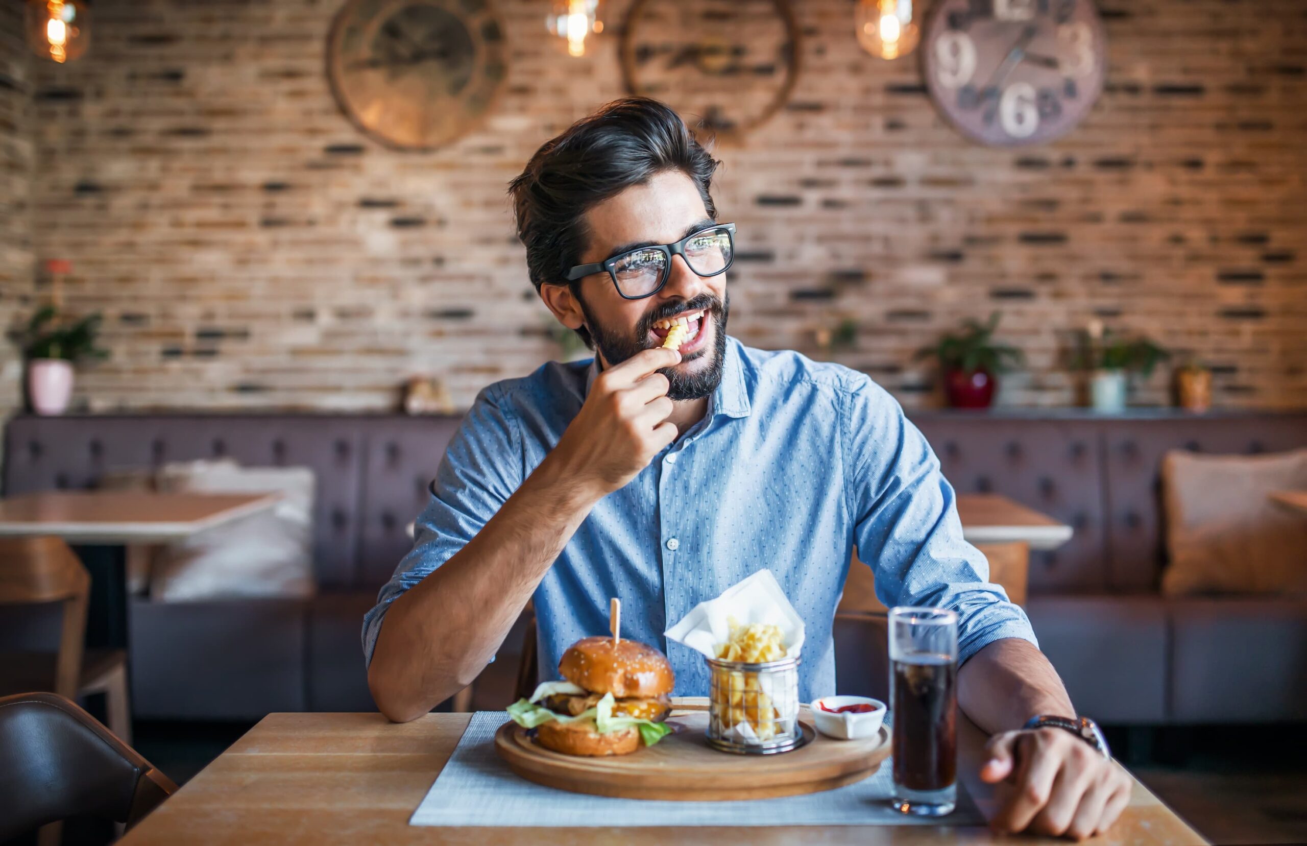 Mann mit Brille sitzt in einem Restaurant und isst Pommes, vor sich ein Burger mit Beilage und ein Getränk, wirkt entspannt und geniesst sein Essen.