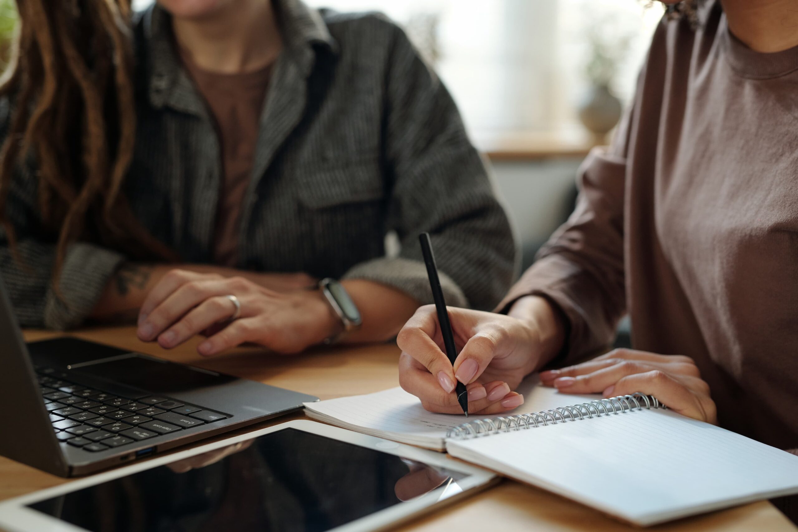 Zwei Personen sitzen an einem Tisch, eine schreibt in ein Notizbuch, während nebenbei ein Laptop und ein Tablet liegen, wirken konzentriert bei der gemeinsamen Arbeit.